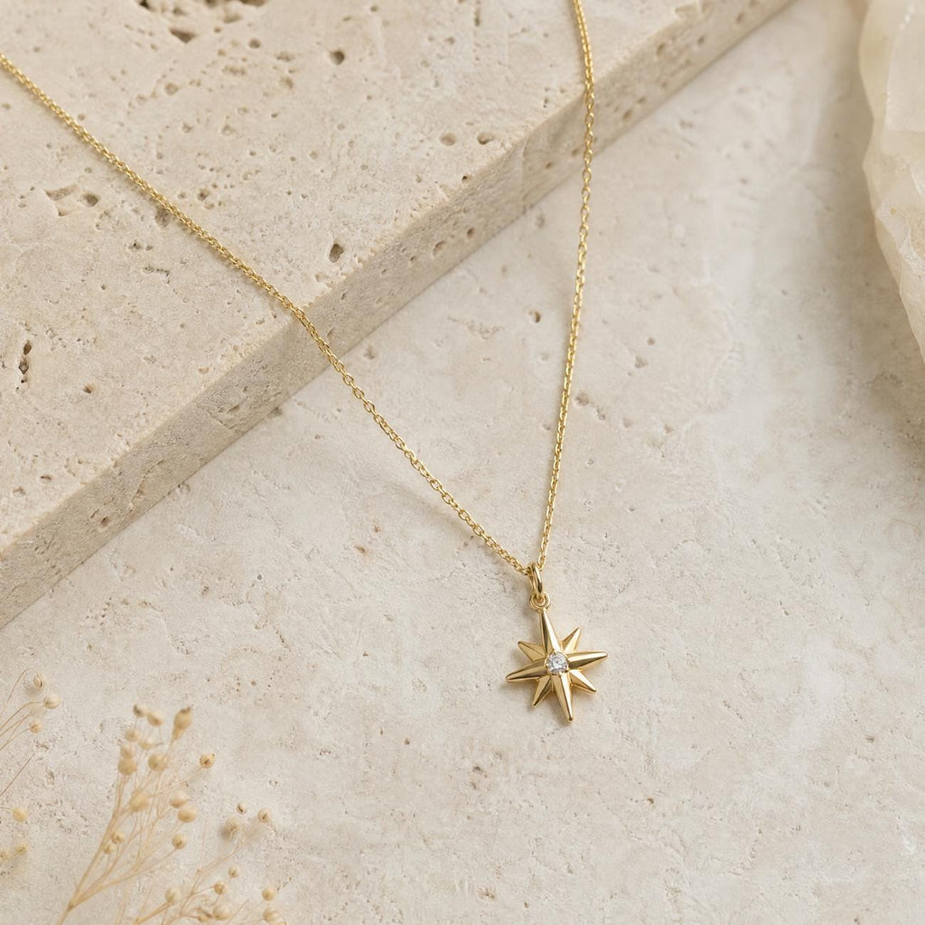 A gold necklace featuring a starburst pendant. The pendant has eight points and a small, clear gemstone at its centre. The chain is fine. The background is a light beige, textured surface.