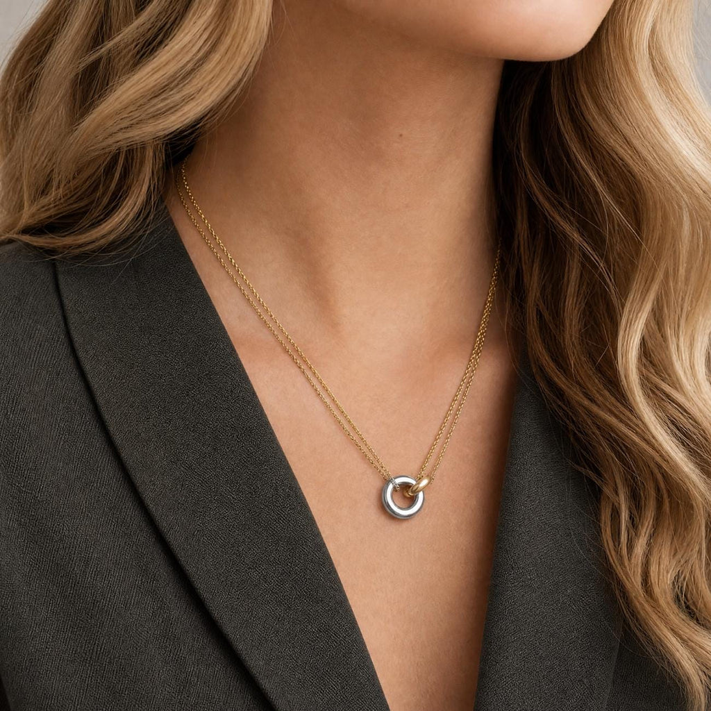 A gold plated double chain necklace featuring a central pendant composed of a small silver-toned ring linked to a gold-toned ring. The necklace rests against the neck, showcasing a design with two distinct metal finishes.