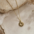 Gold plated necklace featuring a delicate chain and a circular pendant with radiating sunburst-like rays. The pendant has a textured, beaded edge design. The metal has a matte finish, and the chain is thin with small, uniform links.