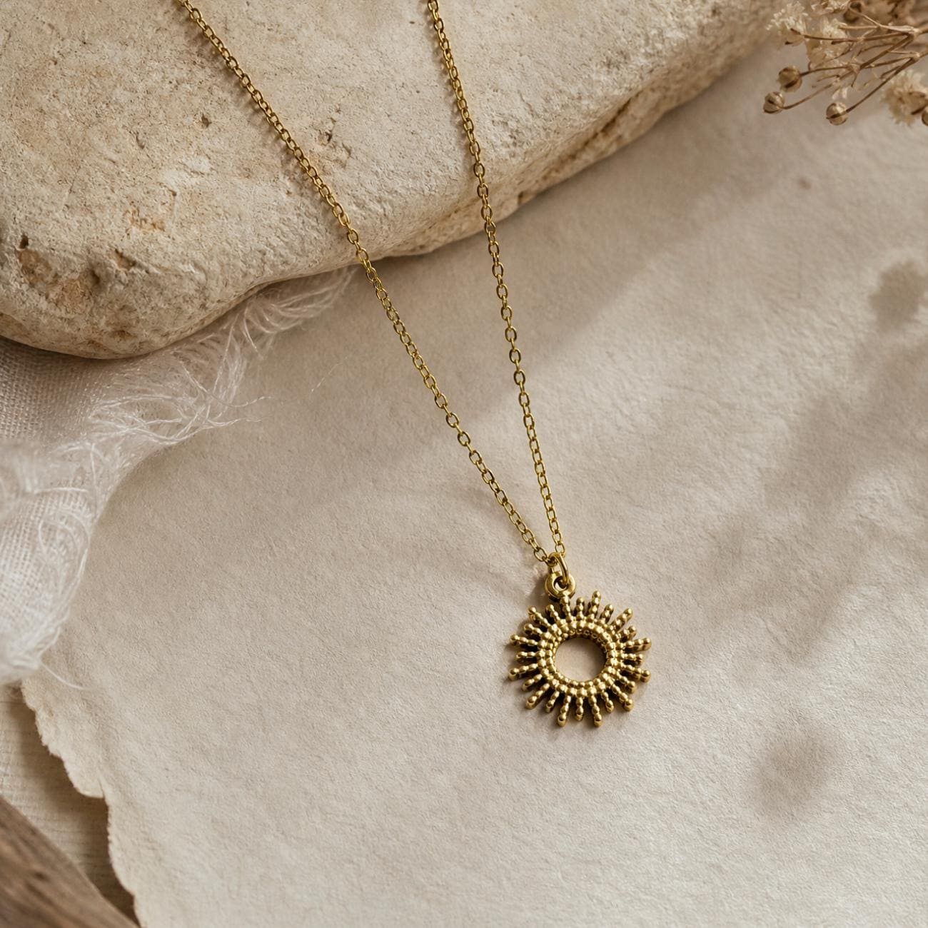 Gold plated necklace featuring a delicate chain and a circular pendant with radiating sunburst-like rays. The pendant has a textured, beaded edge design. The metal has a matte finish, and the chain is thin with small, uniform links.