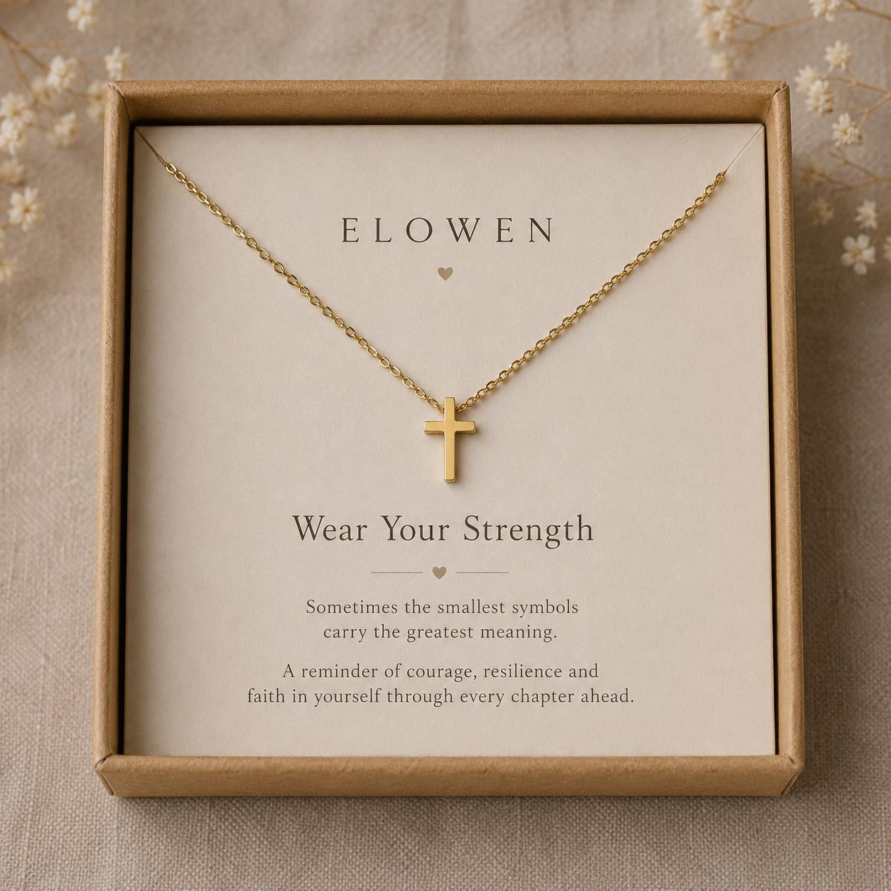 Gold plated necklace with a small cross pendant displayed on a card inside a brown box. The card features the text ELOWEN, Wear Your Strength, and a message about symbols, courage, resilience, and faith in yourself through every chapter.