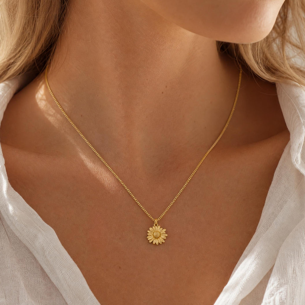 Gold plated necklace featuring a delicate chain and a small, circular pendant shaped like a daisy flower with textured petals and a central stamen. The metal has a matte finish, and the pendant hangs centrally from the thin chain.