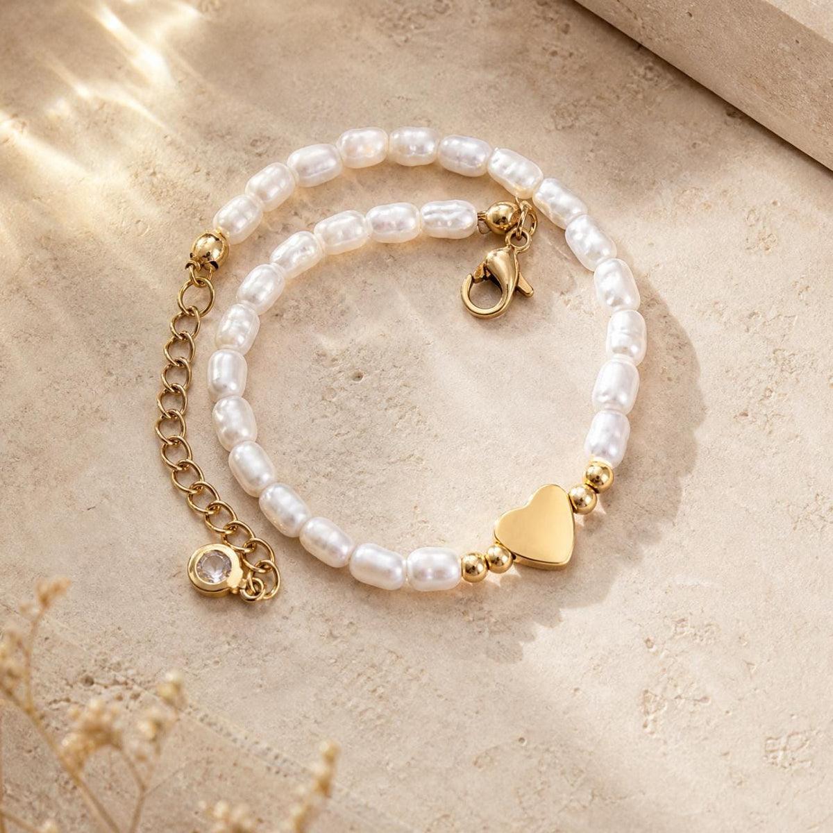 A bracelet featuring white, oblong-shaped pearls arranged in a circular pattern. The piece includes a gold-toned metal heart charm, a gold-toned lobster clasp, and an adjustable chain extension finished with a small, clear crystal bead.