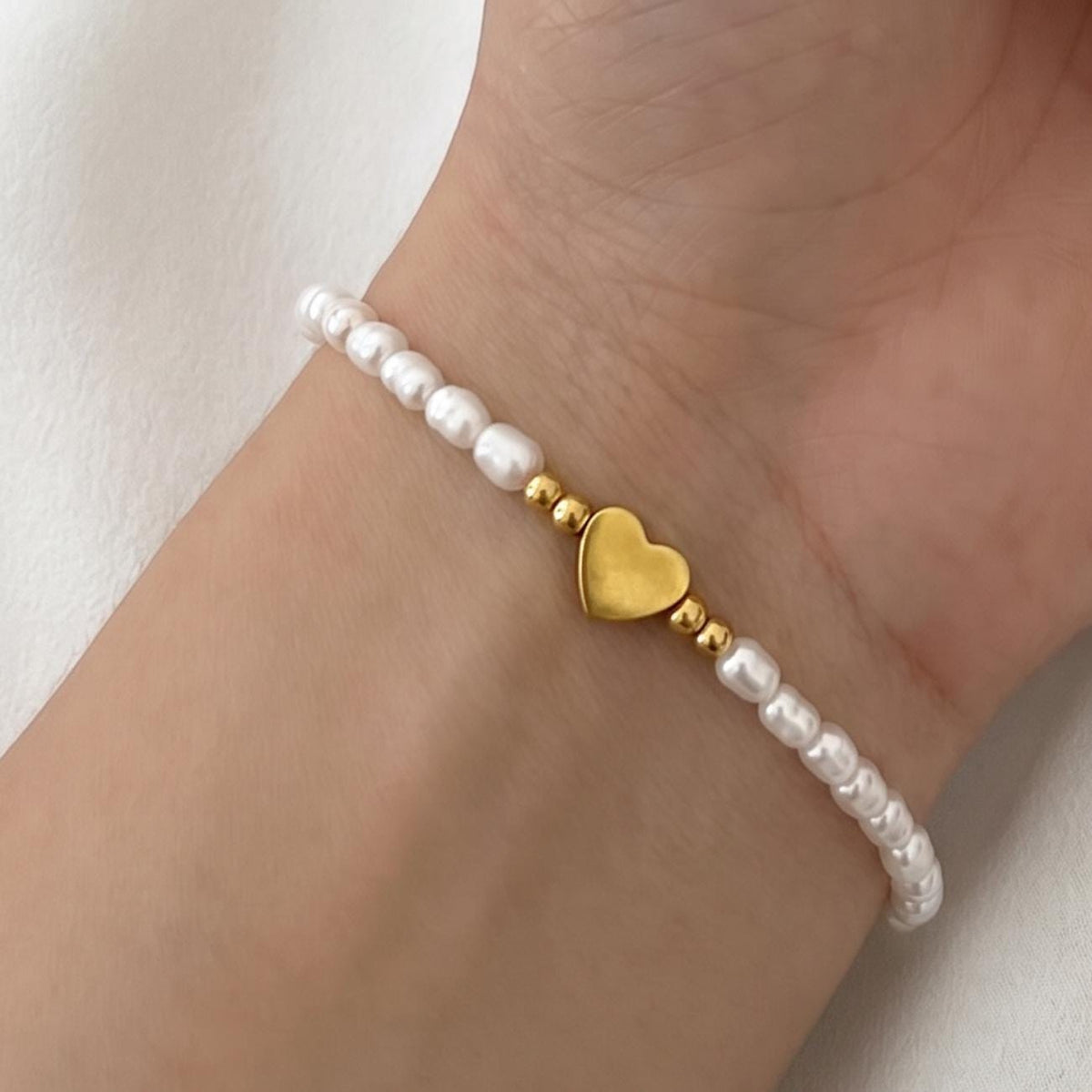 A bracelet featuring a strand of small, white, irregular-shaped imitation pearls. A single, smooth, gold-toned heart charm is positioned at the centre, flanked by two small, round, gold-toned metal beads on each side of the heart.