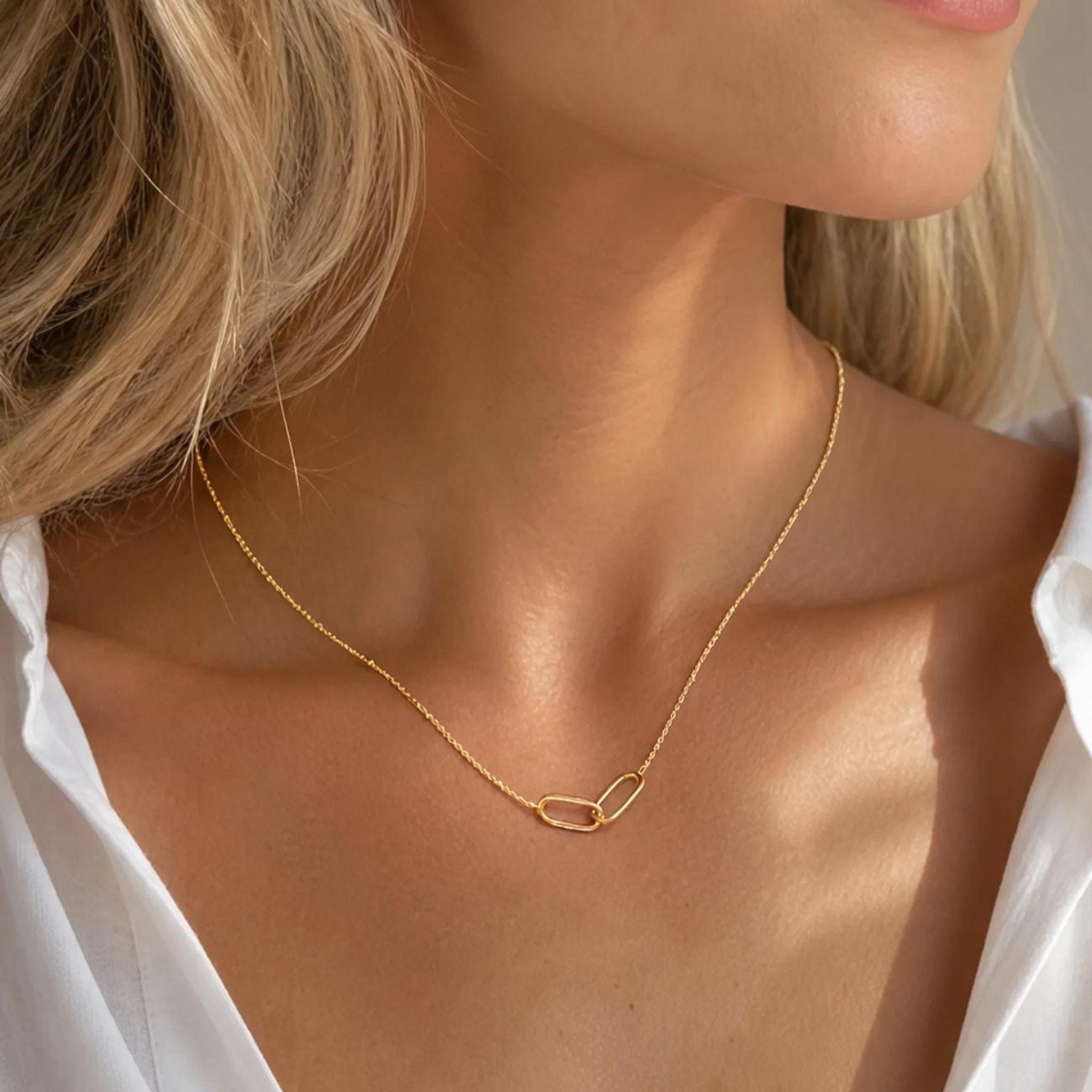 A gold interlocking link necklace with a delicate chain is worn around a person's neck, visible against a white collared shirt.