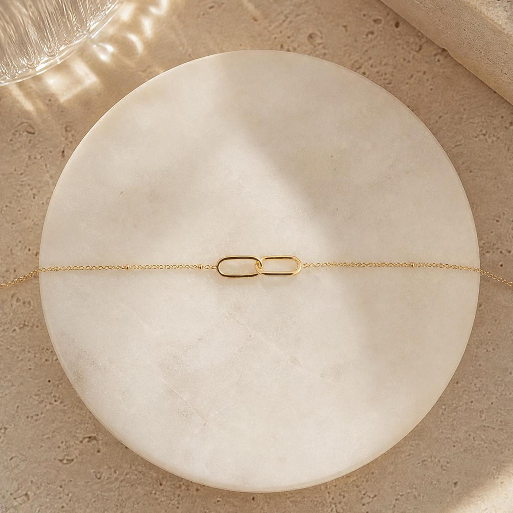 A gold interlocking link bracelet with a delicate chain is laid flat on a round, white marble-like surface.