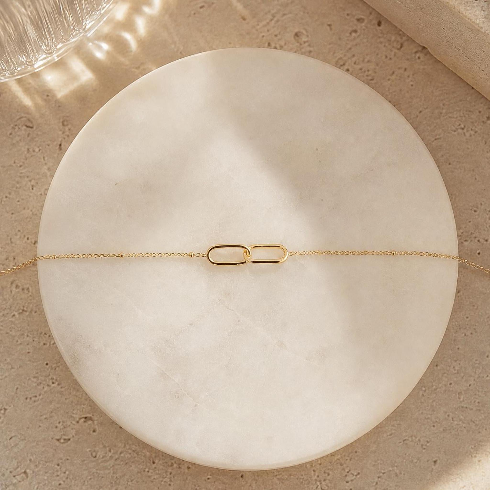 A gold interlocking link bracelet with a delicate chain is laid flat on a round, white marble-like surface.