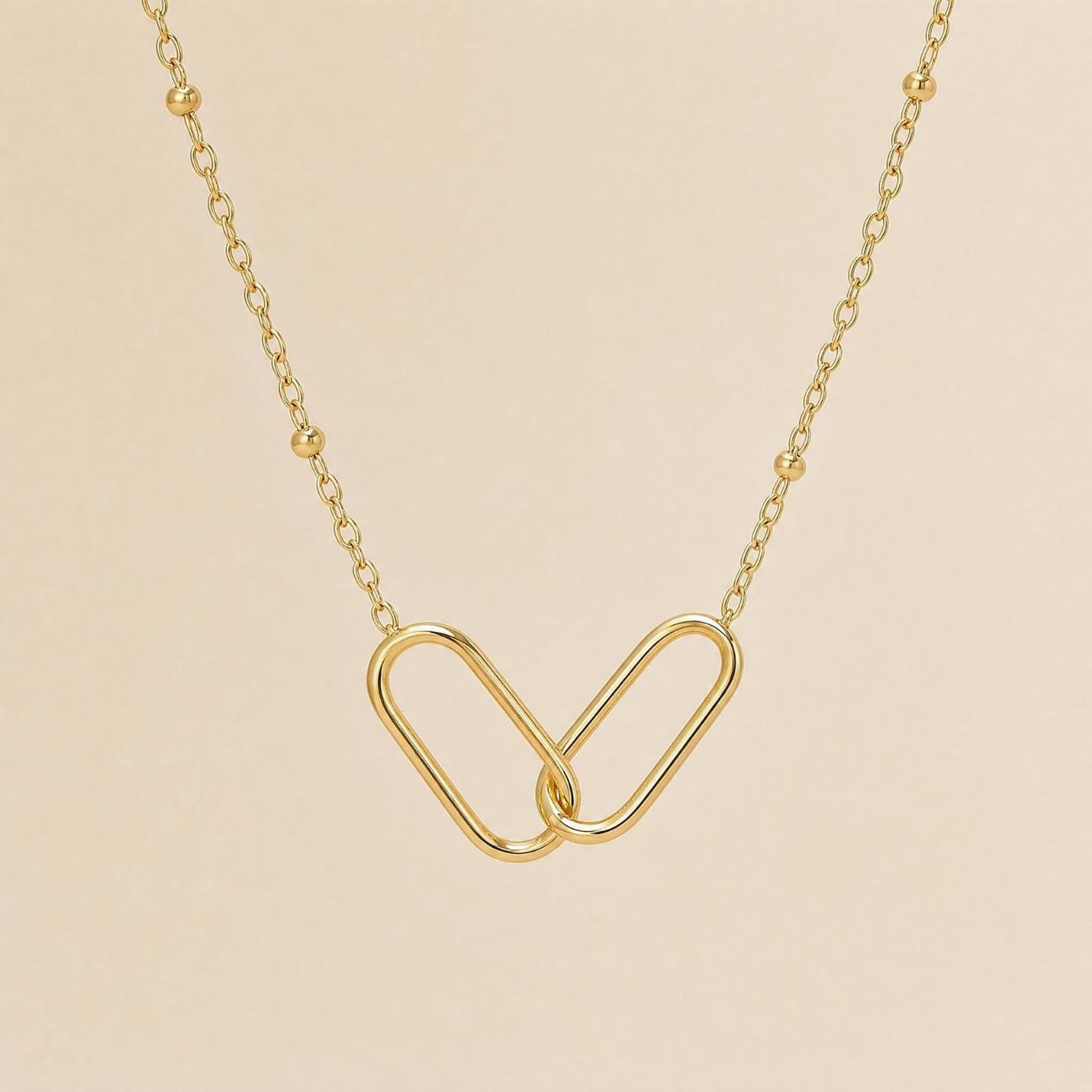 A close-up, overhead view of a gold interlocking link necklace with a beaded chain on a light beige background.