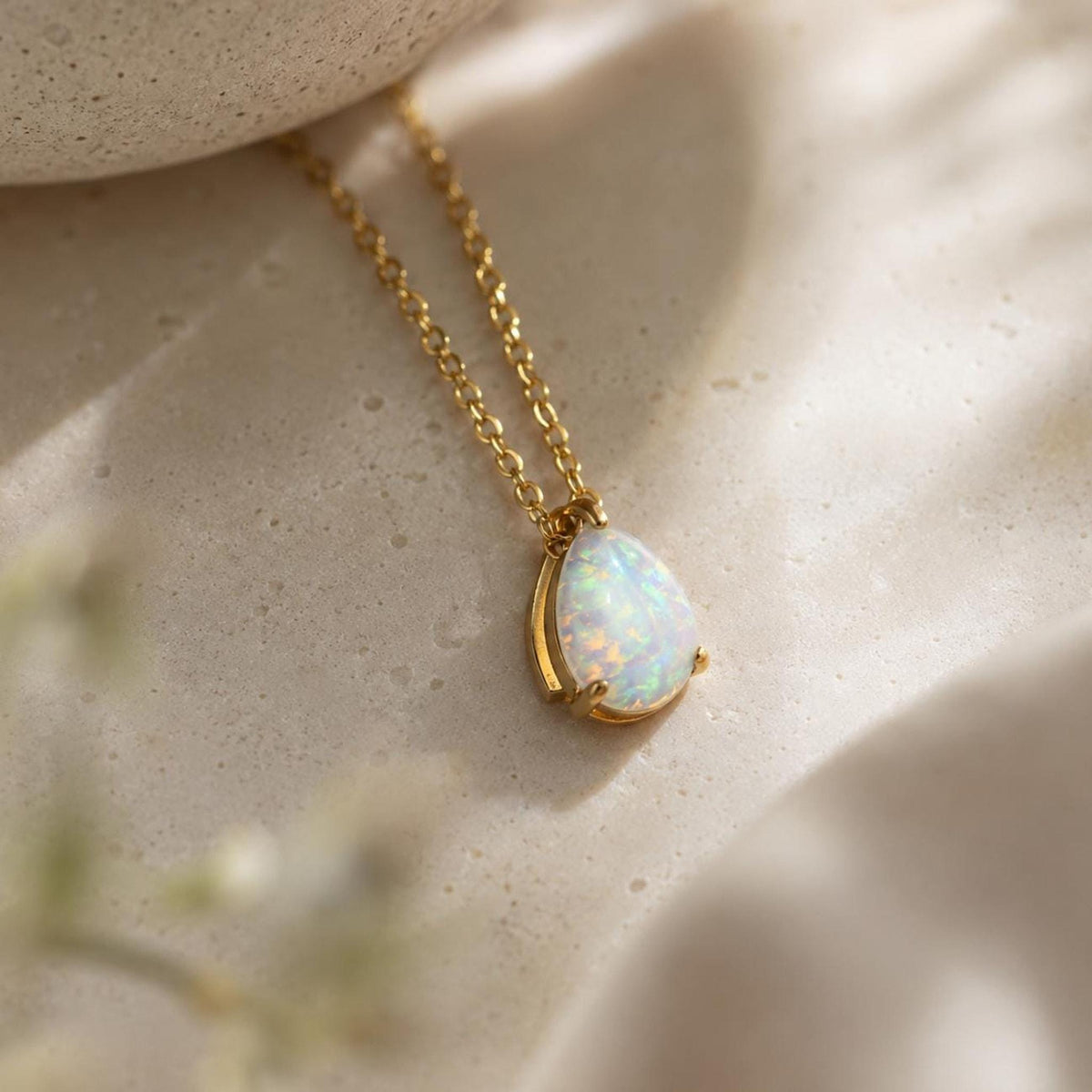 A gold chain necklace with a small, teardrop-shaped white opal pendant is displayed on a light-colored stone surface.