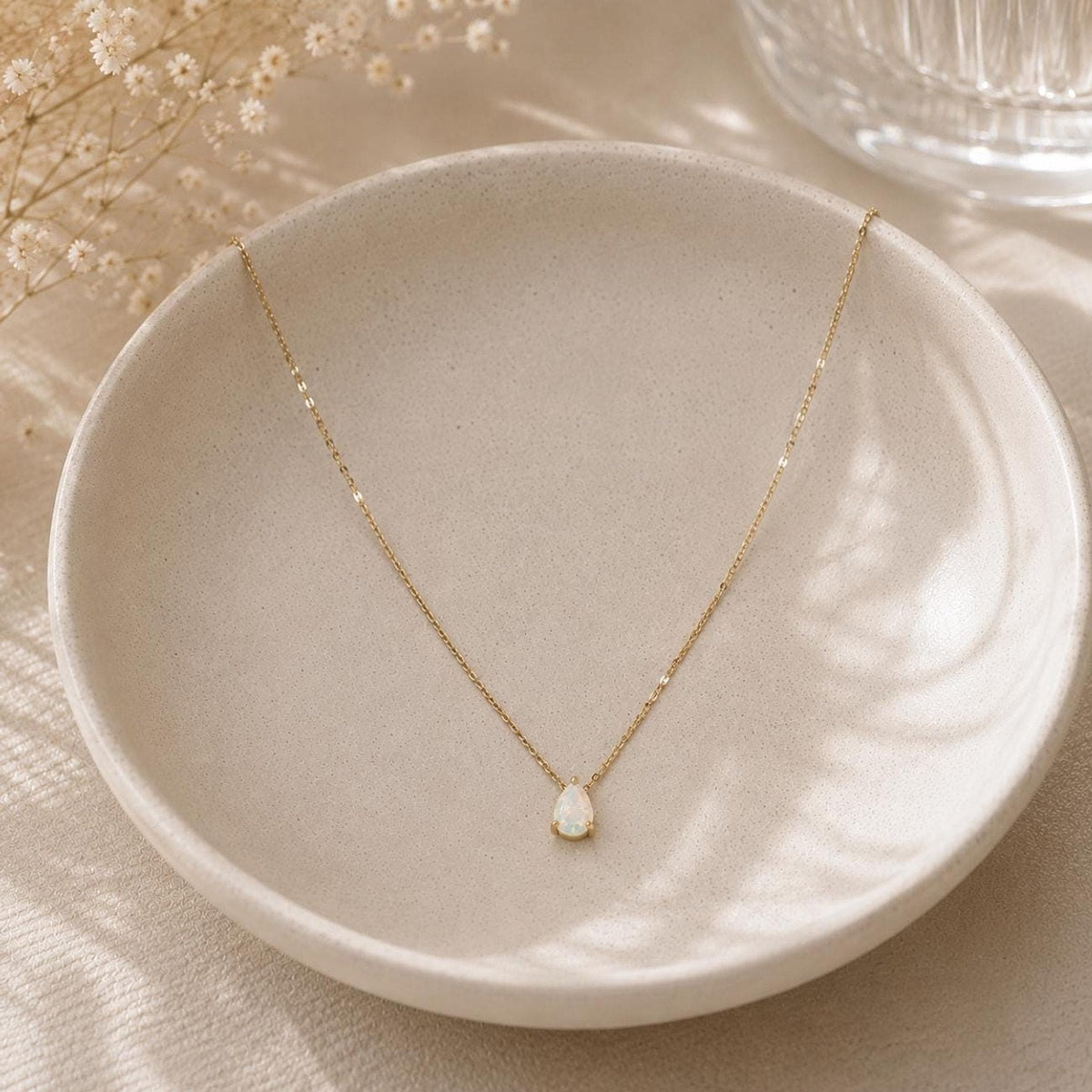 A gold chain necklace with a small, teardrop-shaped white opal pendant rests inside a shallow, light-colored ceramic bowl.