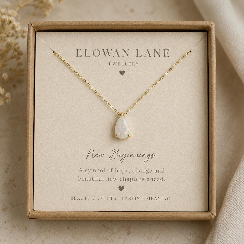 A gold chain necklace with a teardrop white opal pendant is displayed on a card inside a brown gift box, with text including "ELOWAN LANE JEWELLERY" and "New Beginnings."