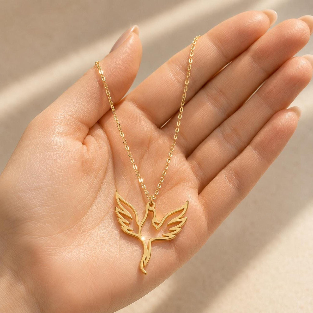 A gold phoenix necklace with a delicate chain and a cutout phoenix pendant, held in the palm of a person's hand.
