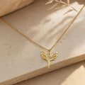 A gold phoenix necklace with a delicate chain and a cutout phoenix pendant, lying on a light beige textured surface with a blurred plant stem in the background.