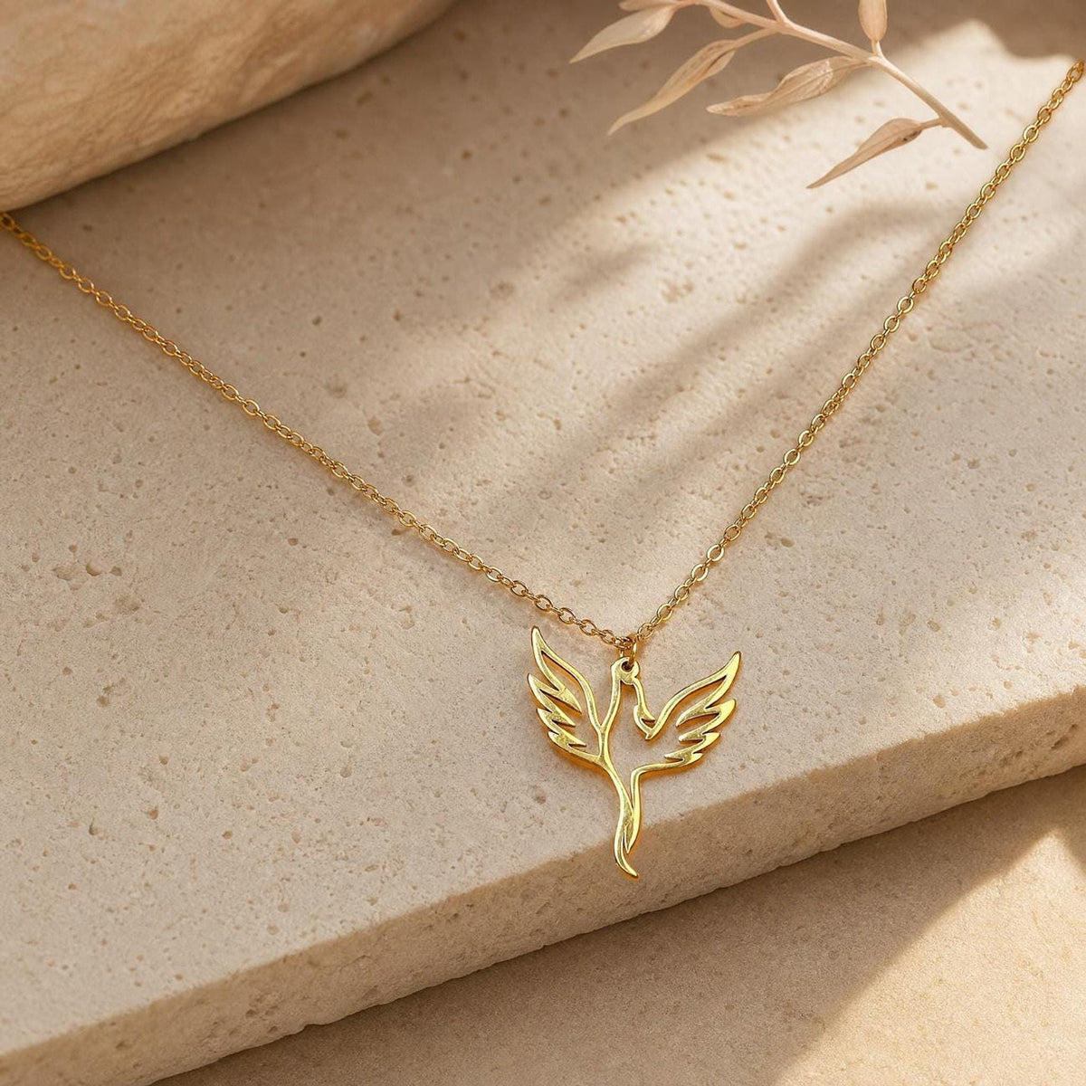 A gold phoenix necklace with a delicate chain and a cutout phoenix pendant, lying on a light beige textured surface with a blurred plant stem in the background.
