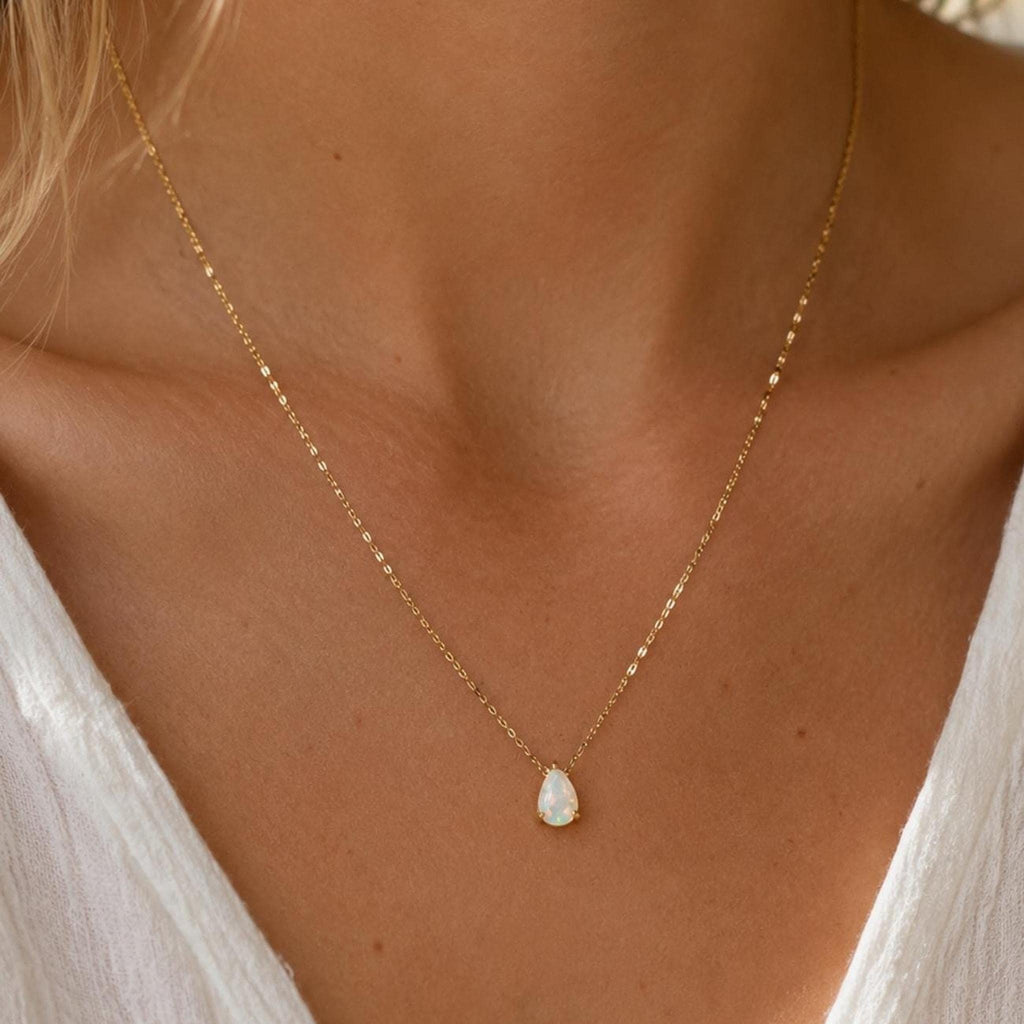 A gold chain necklace with a small, teardrop-shaped white opal pendant hangs on a person wearing a white shirt.