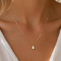 A gold chain necklace with a small, teardrop-shaped white opal pendant hangs on a person wearing a white shirt.