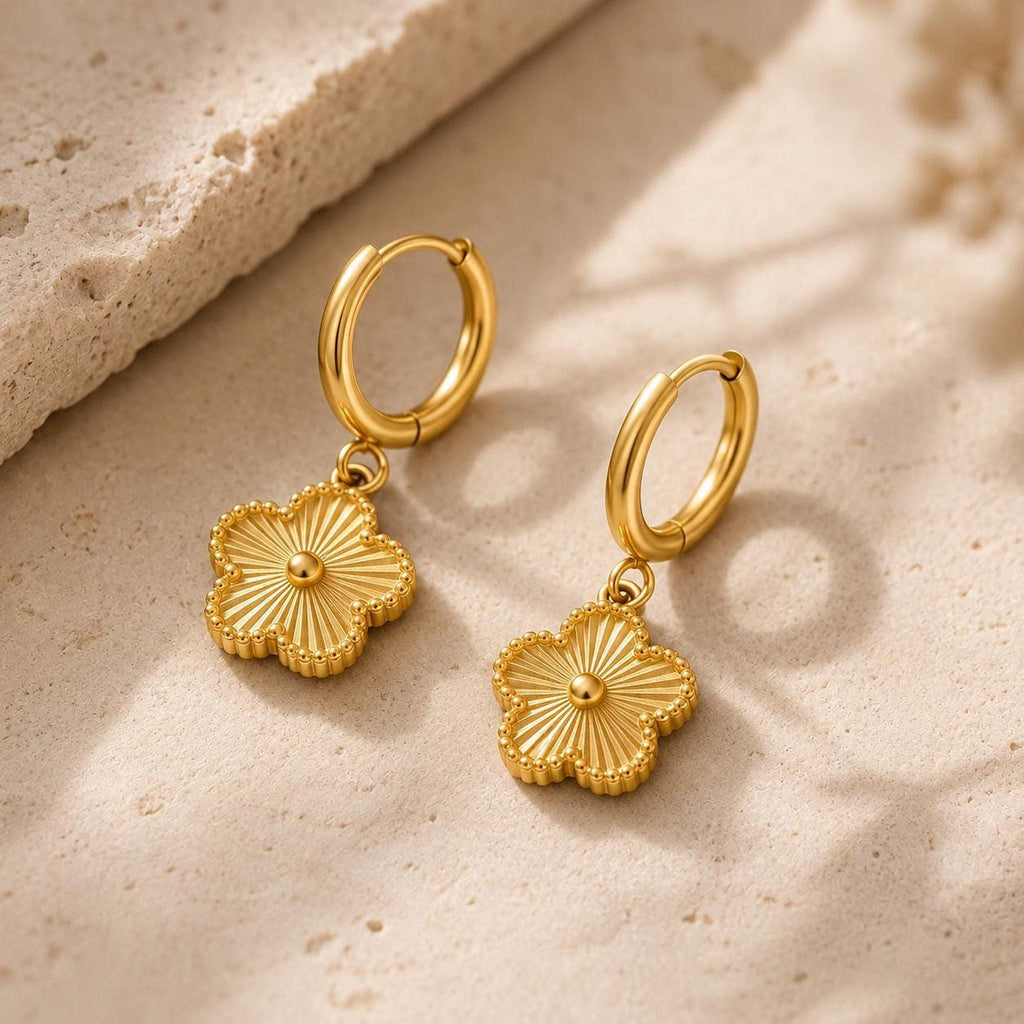 Two gold huggie hoop earrings with textured flower charms resting on a textured beige stone surface.