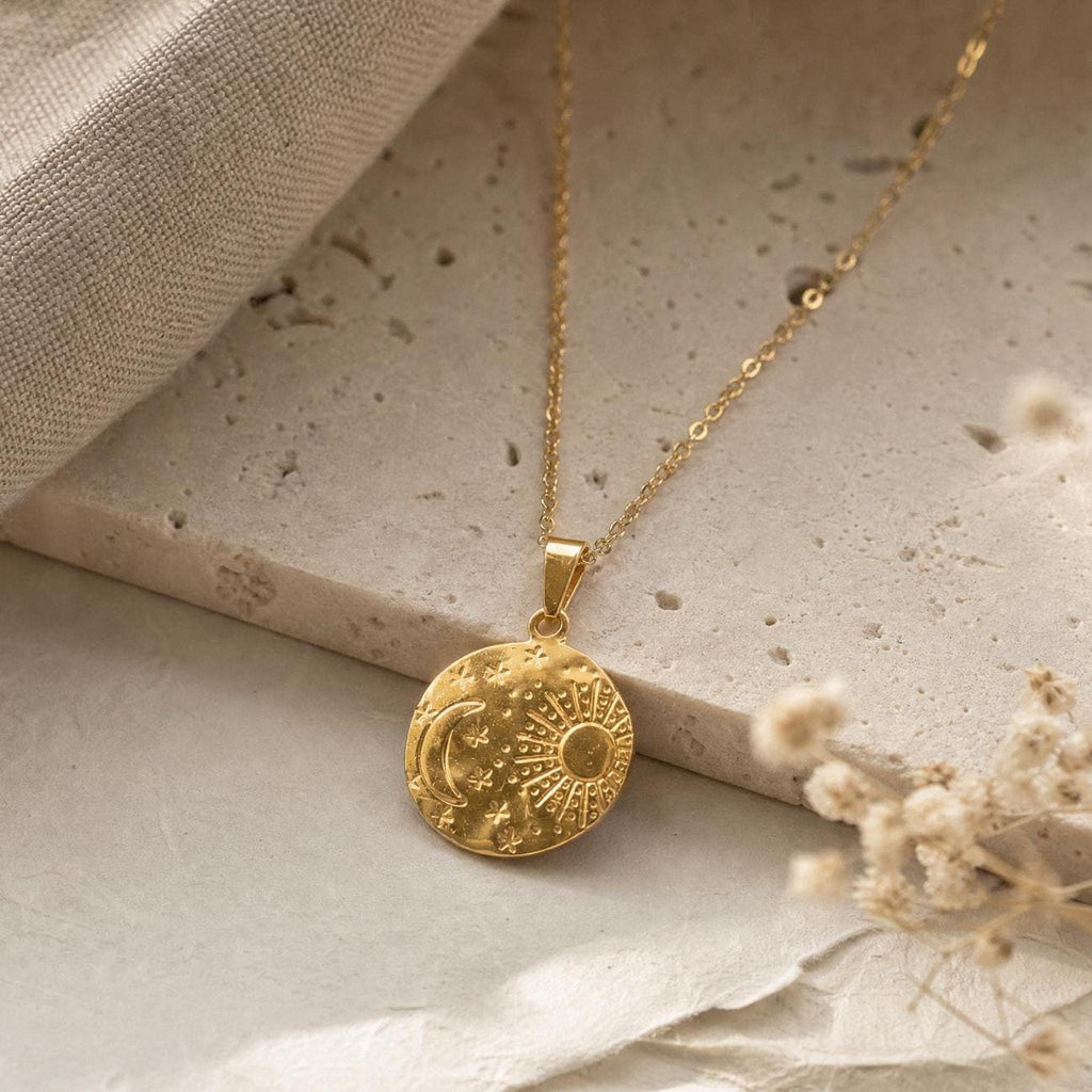 A gold chain necklace with a circular pendant featuring an engraved sun, moon, and stars rests on textured stone.