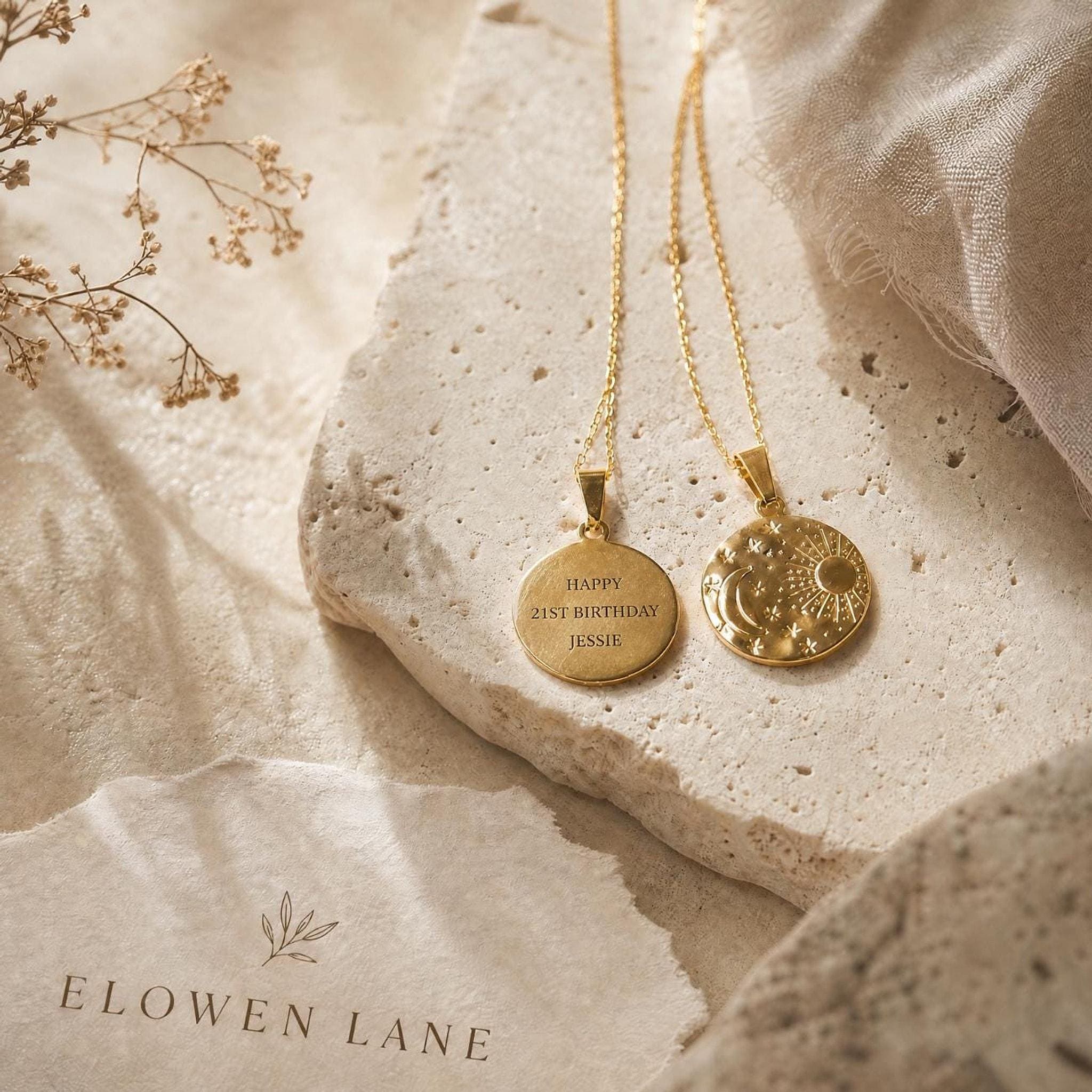 Two gold disc pendant necklaces rest on a light textured stone surface alongside a brand label for Elowen Lane.