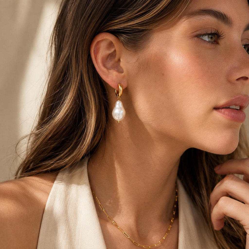 A close-up side profile of a woman wearing a gold huggie hoop earring with a baroque pearl drop and a matching gold chain necklace.