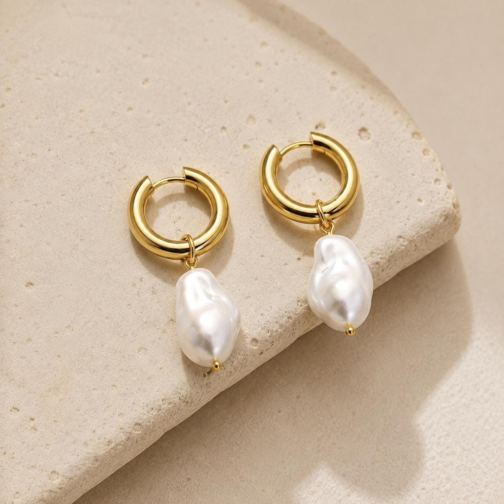 Two gold hoop earrings with hanging baroque freshwater pearls resting on a textured beige surface.