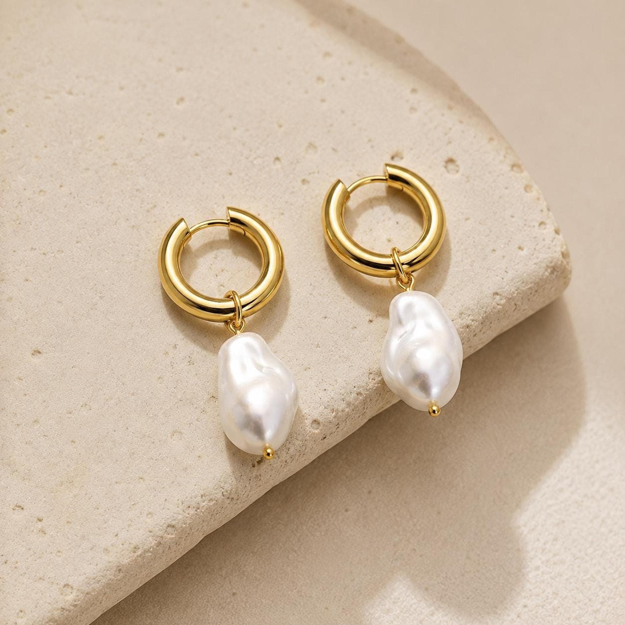 Two gold hoop earrings with hanging baroque freshwater pearls resting on a textured beige surface.